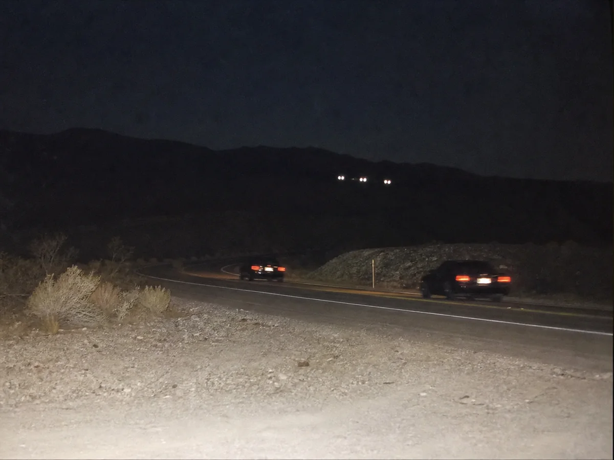 Two dark sedans on a desert road with three pairs of white lights low near a distant ridge