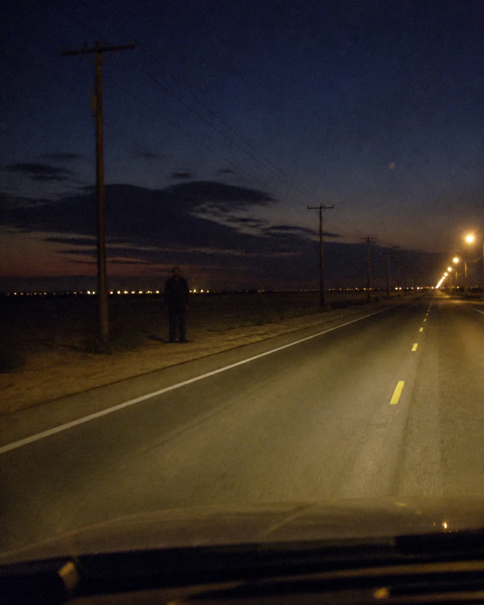 An unidentified adult standing far off the shoulder of a two-lane road after dusk